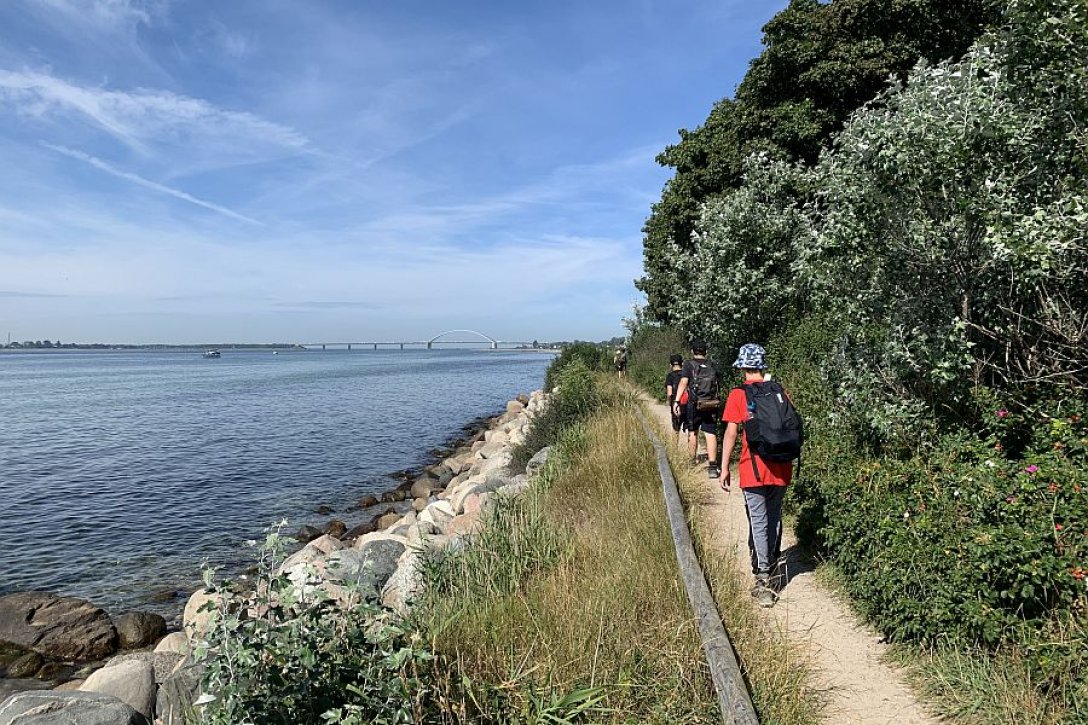 Jugendliche mit Rucksäcken gehen schmalen Wanderweg am Wasser entlang 