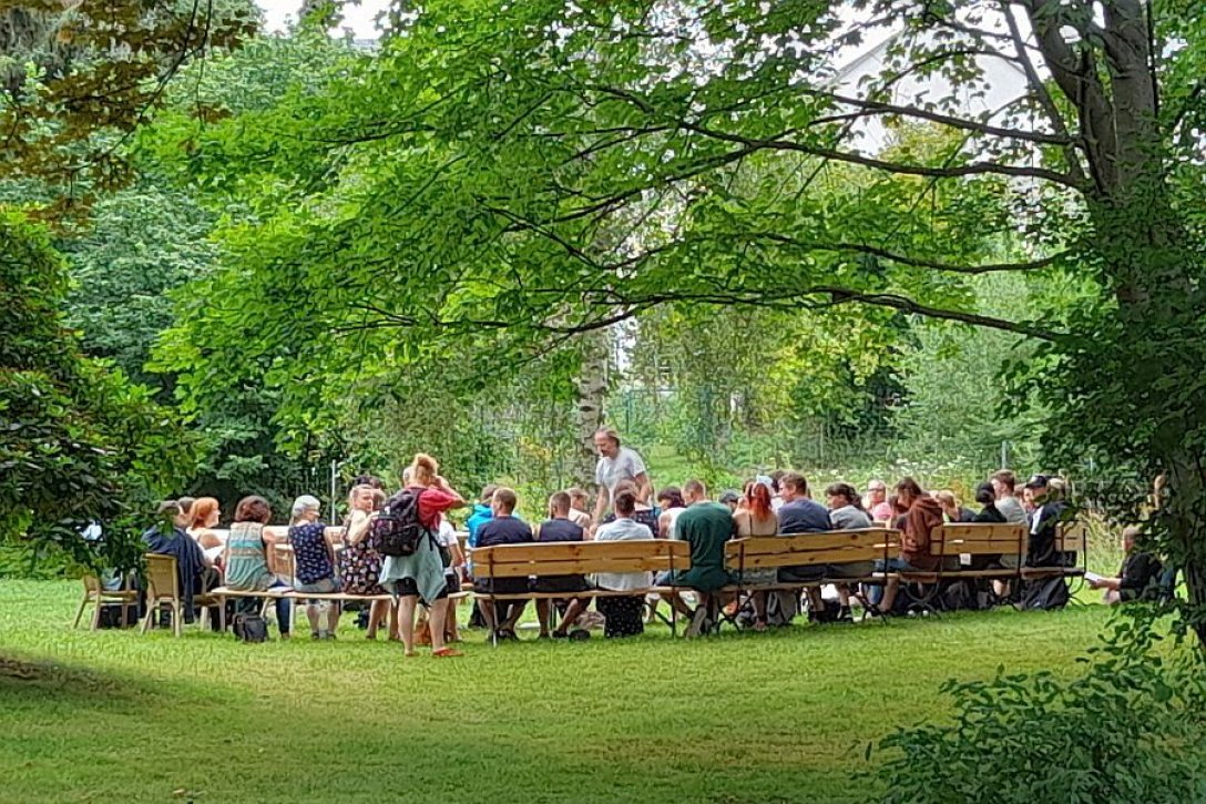 Große Gruppe von jungen Menschen sitzt mit Betreuern auf Bänken auf einer Wiese